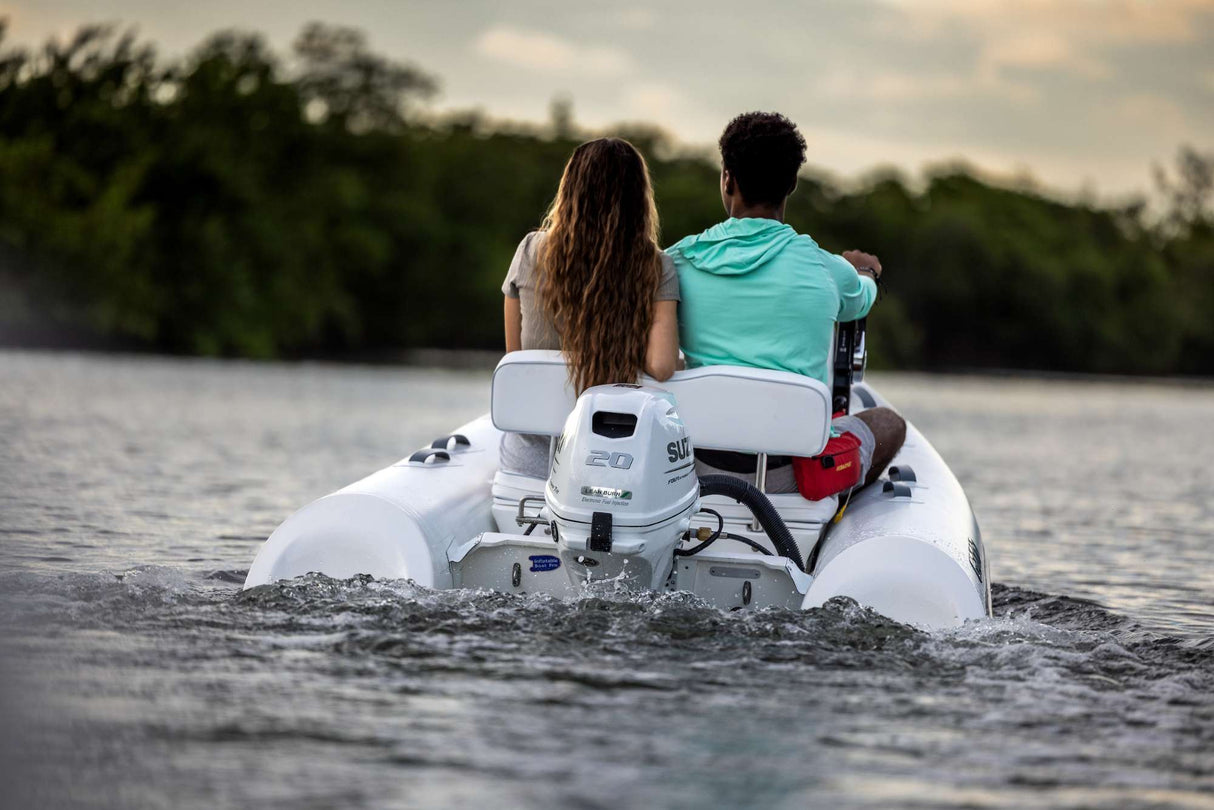 "Couple riding a white inflatable boat powered by a Suzuki 20 HP outboard motor on calm water near a tree-lined shoreline.
