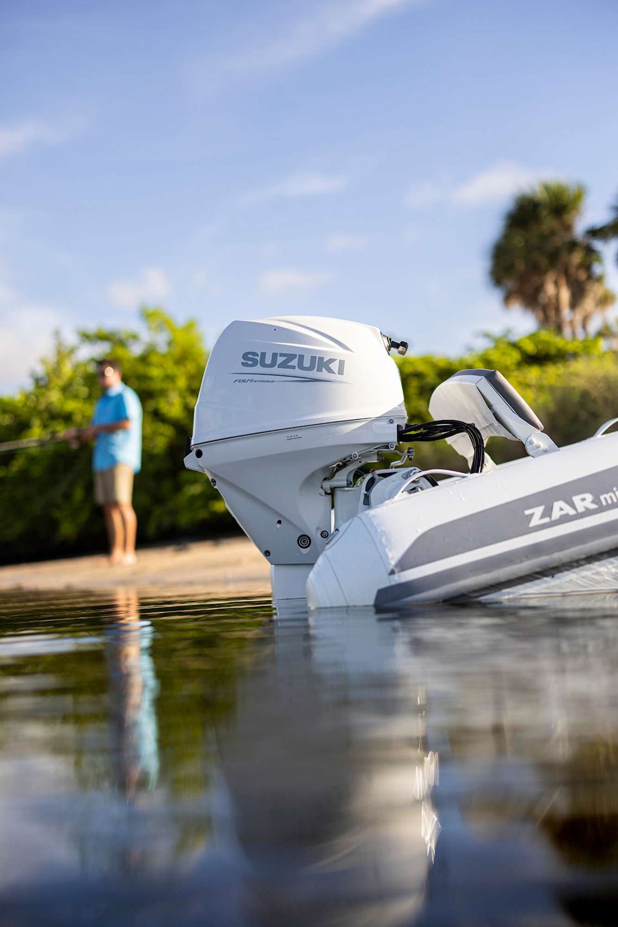 Close-up of a white Suzuki outboard motor on a ZAR mini inflatable boat, with a man fishing from the shore in the background under a sunny sky.