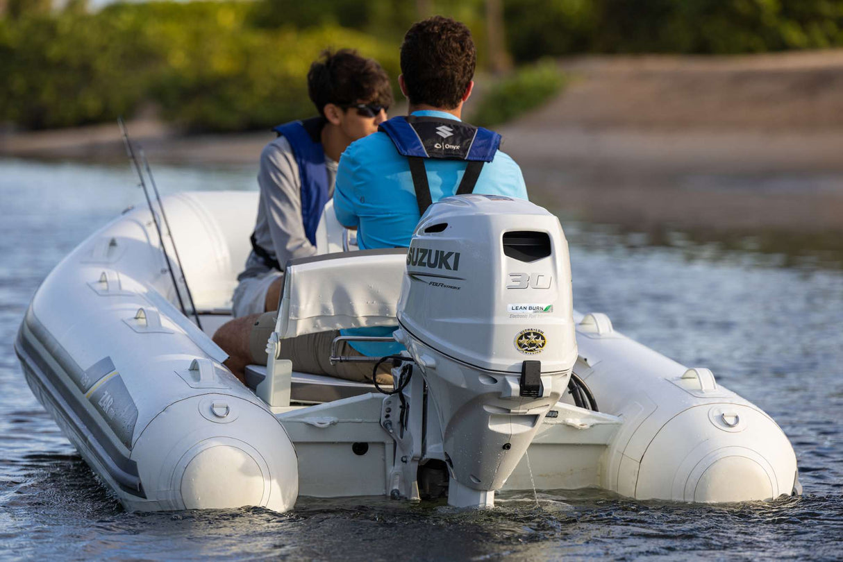 Two men in life jackets navigating a ZAR Mini inflatable boat powered by a Suzuki 30 HP outboard motor on calm water near a shoreline.