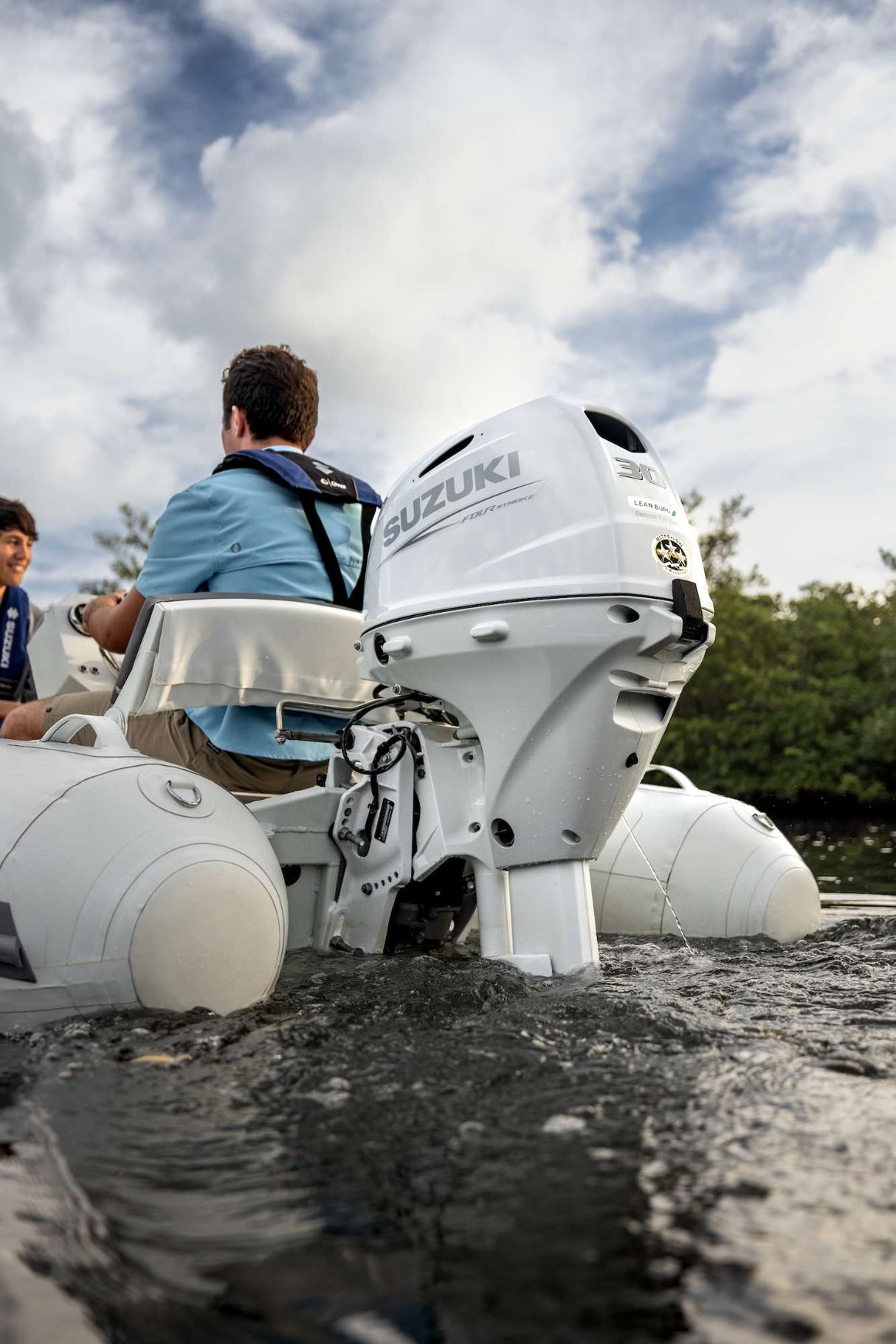 Two people riding a white inflatable boat powered by a Suzuki 30 HP four-stroke outboard motor on calm water under cloudy skies.